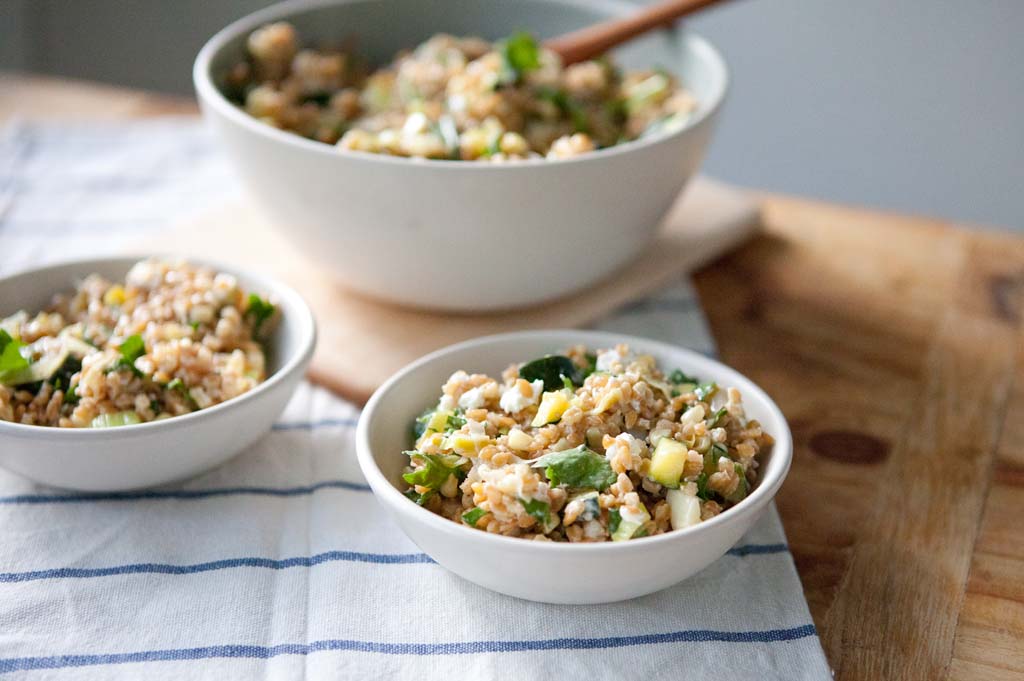 Two small bowls and a large bowl of wheat berry salad served on a striped cloth and wooden board