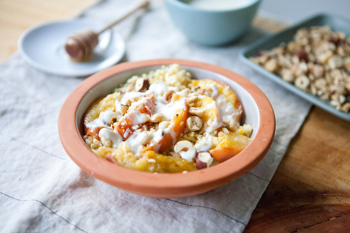 Gluten free caramelized peach millet breakfast bowl topped with yogurt and chopped hazelnuts on a linen napkin, honey dipper and granola in the background