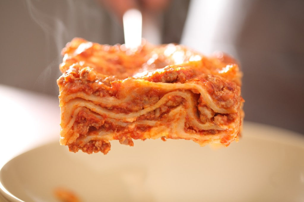 Slice of gluten-free lasagna bolognese lifted above a plate, showing layered pasta, meat ragu, melted cheese, and rising steam