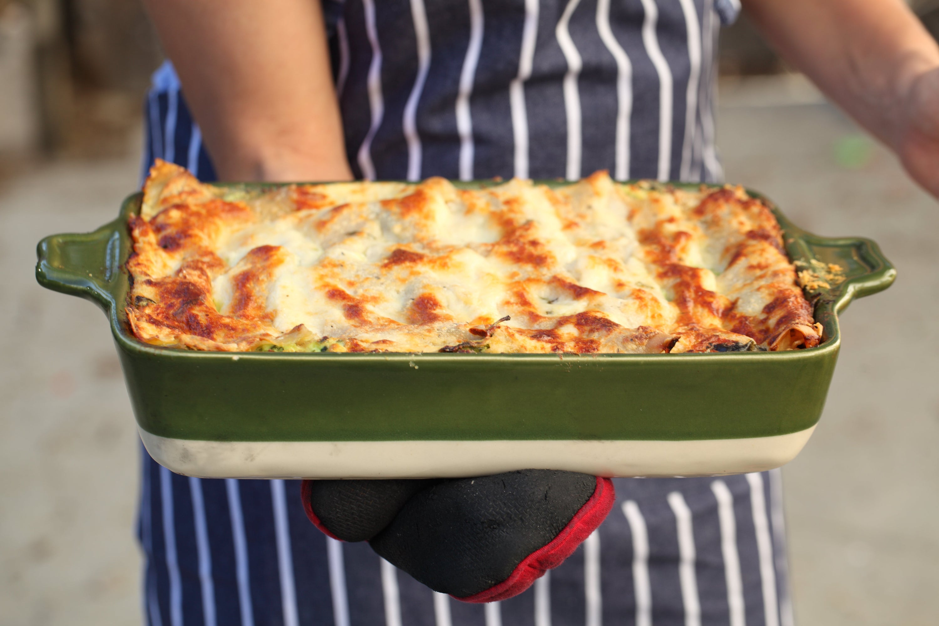 Baked gluten free vegetarian lasagna in a green ceramic baking dish held by a person in a striped apron wearing an oven mitt