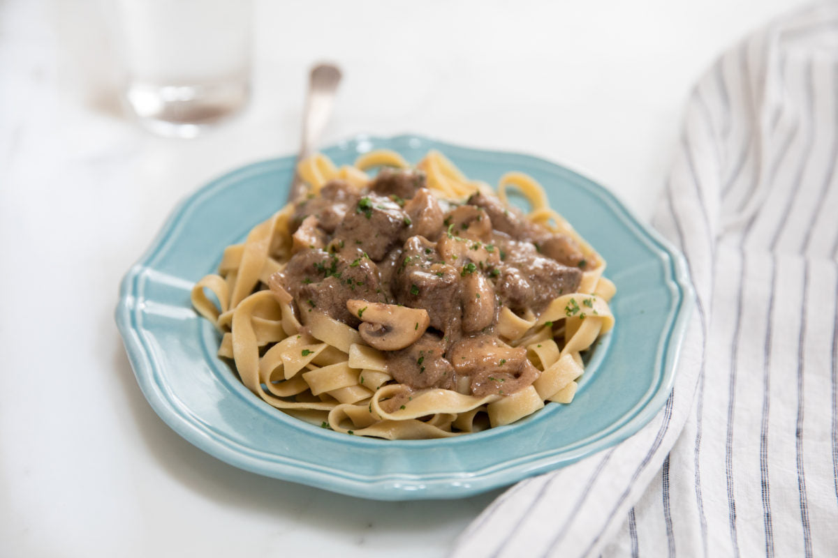 Gluten-free beef stroganoff served over tagliatelle on a blue ceramic plate with a striped napkin nearby