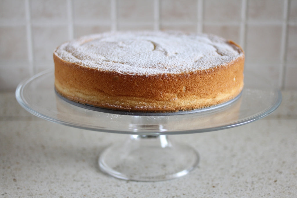 Side view of Gluten Free sweet potato sponge cake dusted with powdered sugar on a glass cake stand against a tiled counter