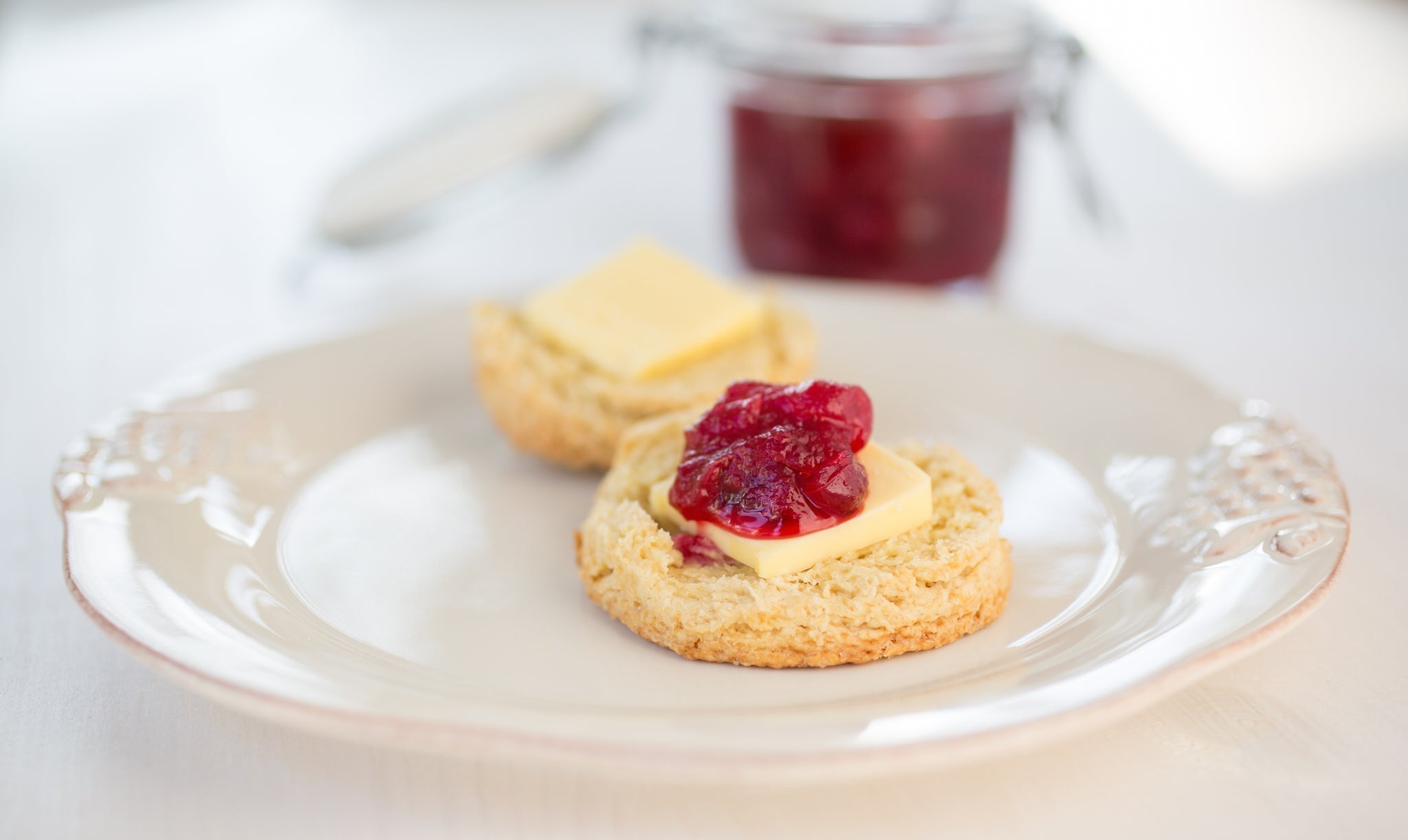 Two split Einkorn buttermilk biscuits on a white plate topped with butter and red jam, jar and butter knife blurred in the background