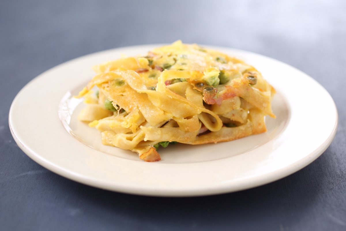 Front view of gluten-free tagliatelle carbonara pasta bake with noodles, melted cheese, peas and bacon on a white plate