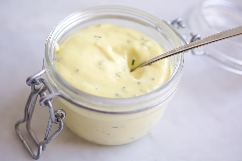 Close-up of a glass jar of homemade herbed olive oil mayonnaise with herb flecks and a spoon on a light surface