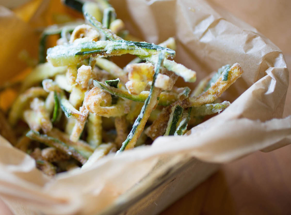 Close-up of thin zucchini strips fried until crispy, served in a parchment-lined paper tray