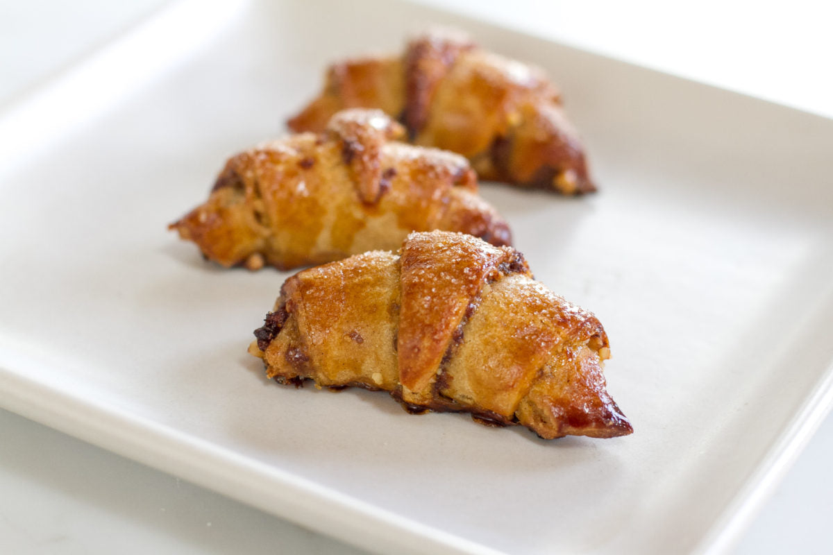 Three-quarter view of three Einkorn rugelach on a white plate showcasing golden-brown flaky crust and sugar-dusted tops