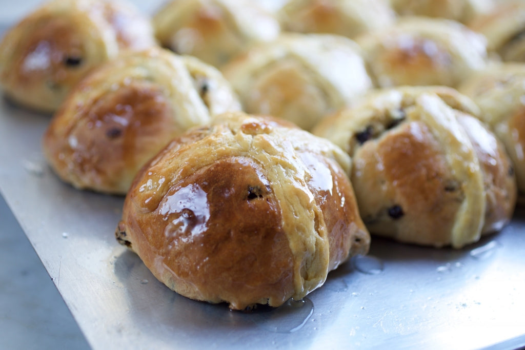 Close up of glazed Einkorn hot cross buns showing golden brown crust, pastry cross and raisins on a metal baking sheet