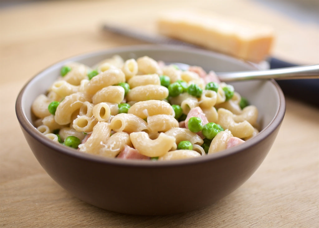 Bowl of creamy elbow pasta with peas and ham in a brown bowl on a wooden table with fork and wedge of cheese in background