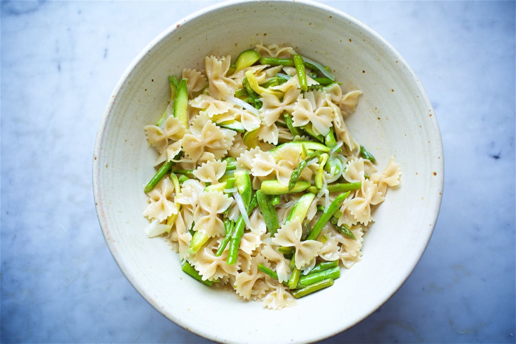 Overhead view of a white ceramic bowl of gluten-free farfalle tossed with asparagus and sliced zucchini on a marble surface