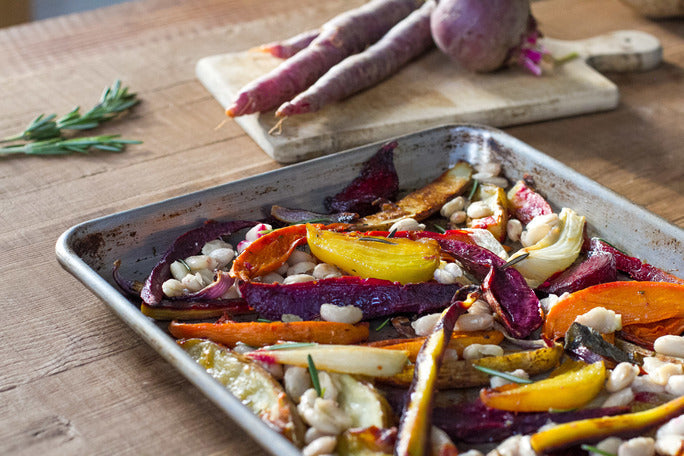 Tray of olive oil roasted root and winter vegetables with white beans and rosemary on a wooden table