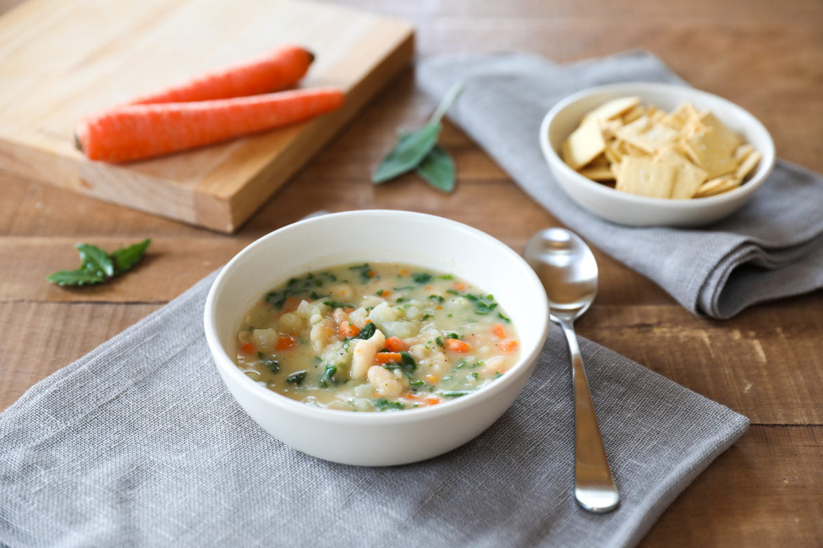 Bowl of cannellini, potato, and vegetable soup on a gray napkin with a spoon and a bowl of crackers on a wooden table