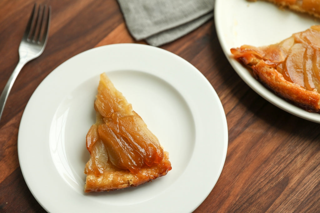 Top-down of a slice of Einkorn apple tarte tatin showcasing caramelized apples on a golden crust on a white plate on wood