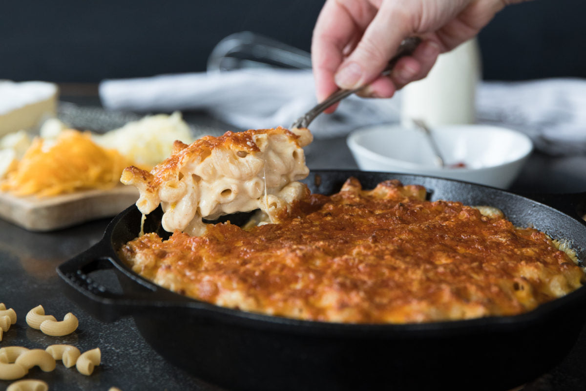 Hand scooping gluten-free mac and cheese from a cast-iron skillet, revealing a golden cheese crust and melted cheese
