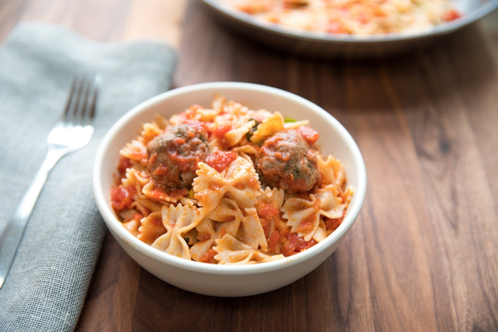 Gluten-free farfalle pasta with marinara and meatballs served in a white bowl with a fork and gray napkin on a wooden table