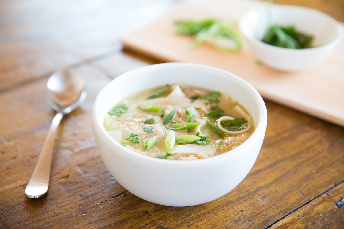 Bowl of Asian chicken soup with gluten-free egg tagliatelle, topped with sliced scallions, on a wooden table with a spoon