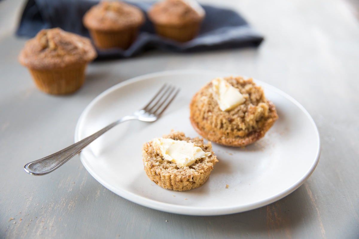 Einkorn spiced apple muffins on a white plate, one split with butter and a fork, extra muffins on a cloth in soft natural light