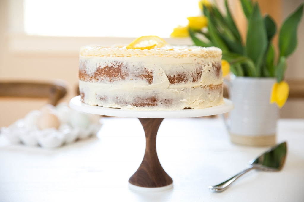 Einkorn lemon layer cake on a marble-and-wood cake stand in a bright kitchen with yellow tulips