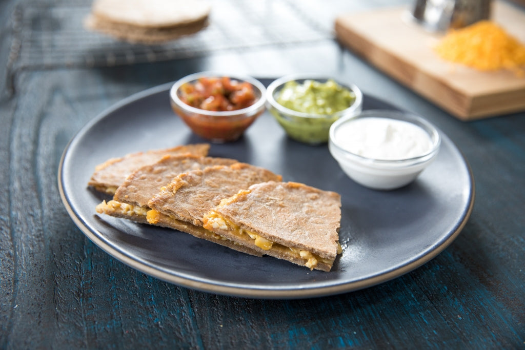Three wedges of whole wheat Einkorn tortilla quesadilla with melted cheese served on a blue plate with salsa, guacamole, and sour cream