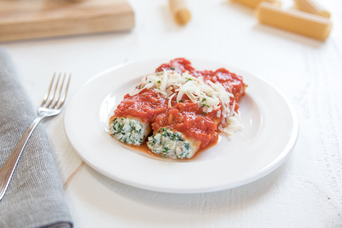 Gluten-free turkey and spinach manicotti on a white plate topped with tomato sauce and shredded cheese, fork and napkin at left