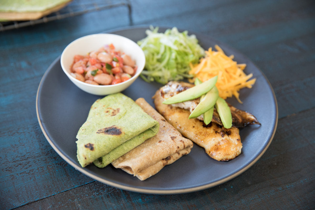 Plate of homemade Einkorn spinach and plain tortillas with grilled fish, avocado, shredded lettuce, cheddar, and beans