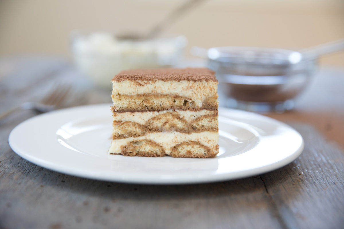 Front view of Einkorn tiramisù slice showing layered cream and espresso-soaked ladyfingers with cocoa dusting on a white plate