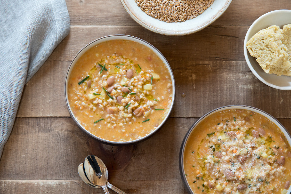 Two bowls of farro and bean soup with einkorn wheat berries, garnished with herbs, on a wooden table with a spoon and a small dish of bread