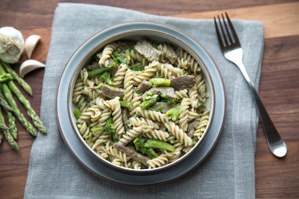 Bowl of Einkorn fusilli tossed with sliced steak, asparagus, and broccoli on a gray napkin with a fork