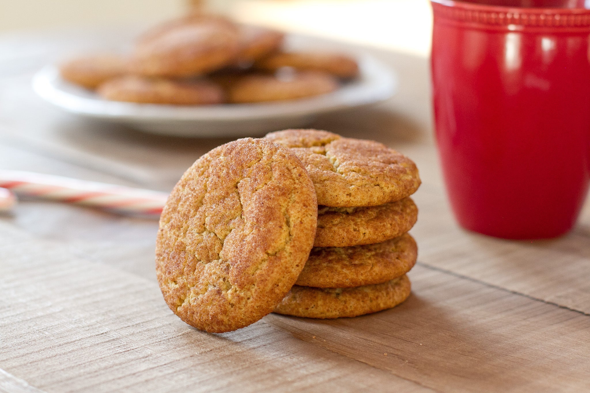 Stack of Einkorn snickerdoodle cookies on a wooden surface with a red mug and a plate blurred in the background