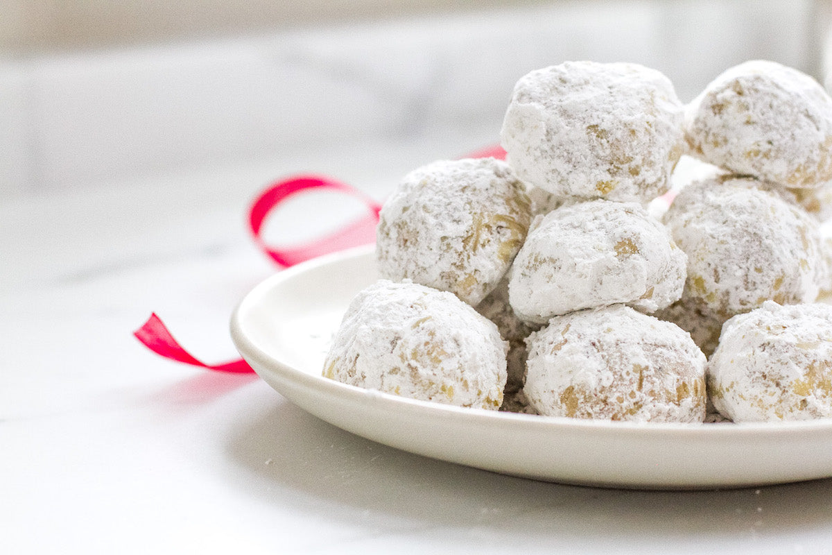 Close-up side view of Einkorn walnut snowball cookies showing powdered sugar coating on a white plate with a red ribbon in the background