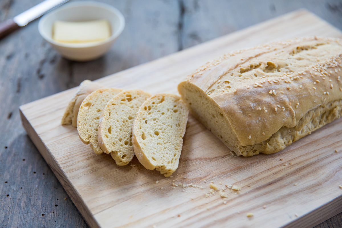 Three-quarter view of Einkorn Italian sourdough loaf showcasing three sliced pieces and sesame seeds on a wooden cutting board with a butter dish in the background