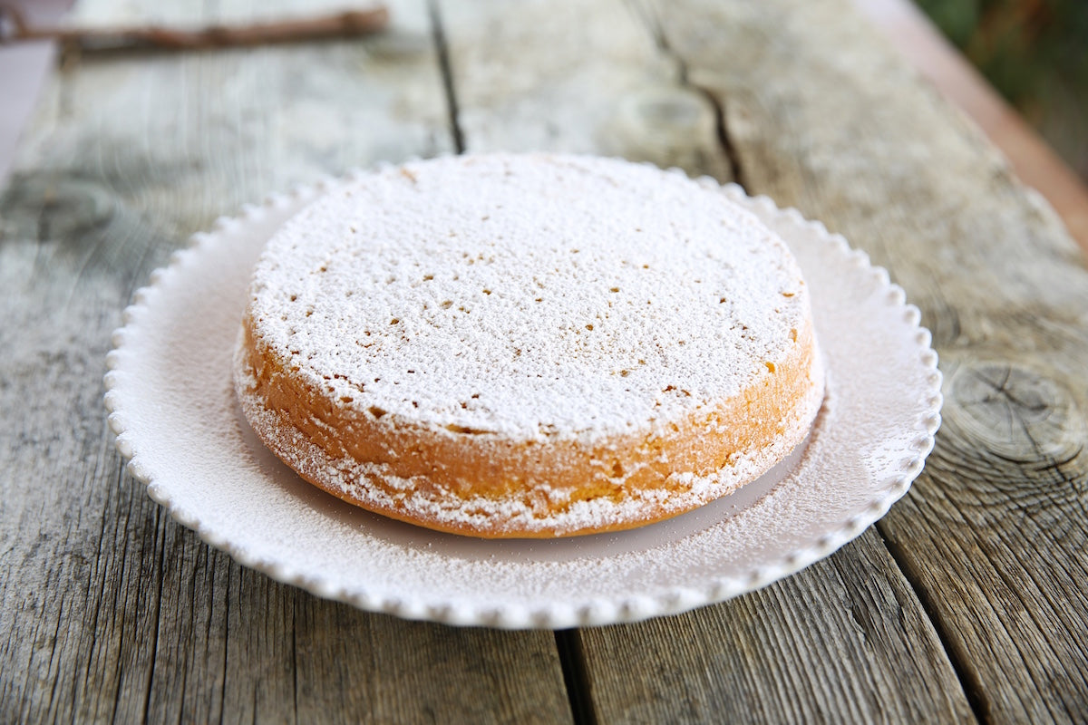 Angled view of Einkorn apple breakfast cake dusted with powdered sugar on scalloped white plate on weathered wooden table