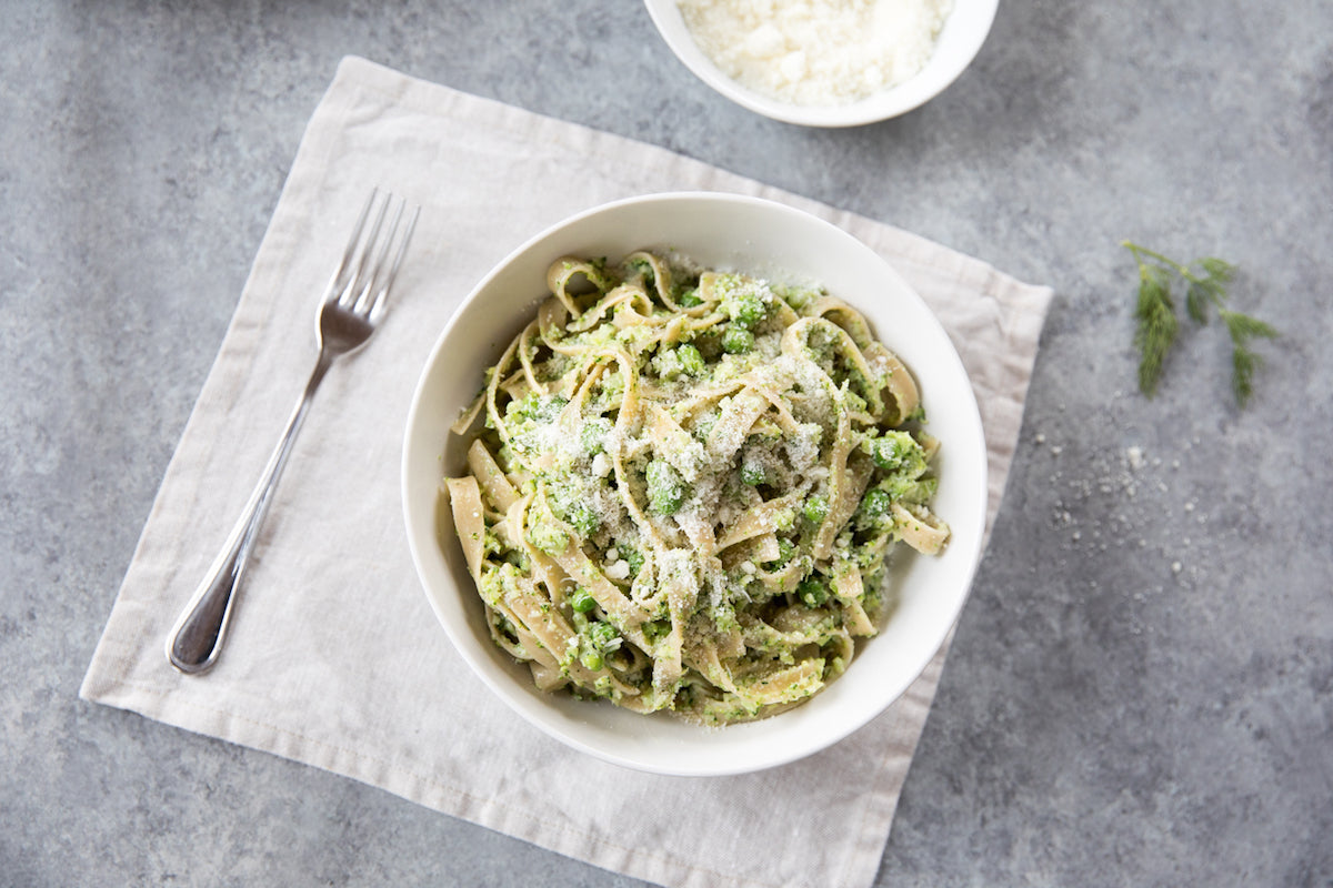 Bowl of creamy tagliatelle tossed with broccoli and peas, sprinkled with grated cheese, on a linen napkin with a fork