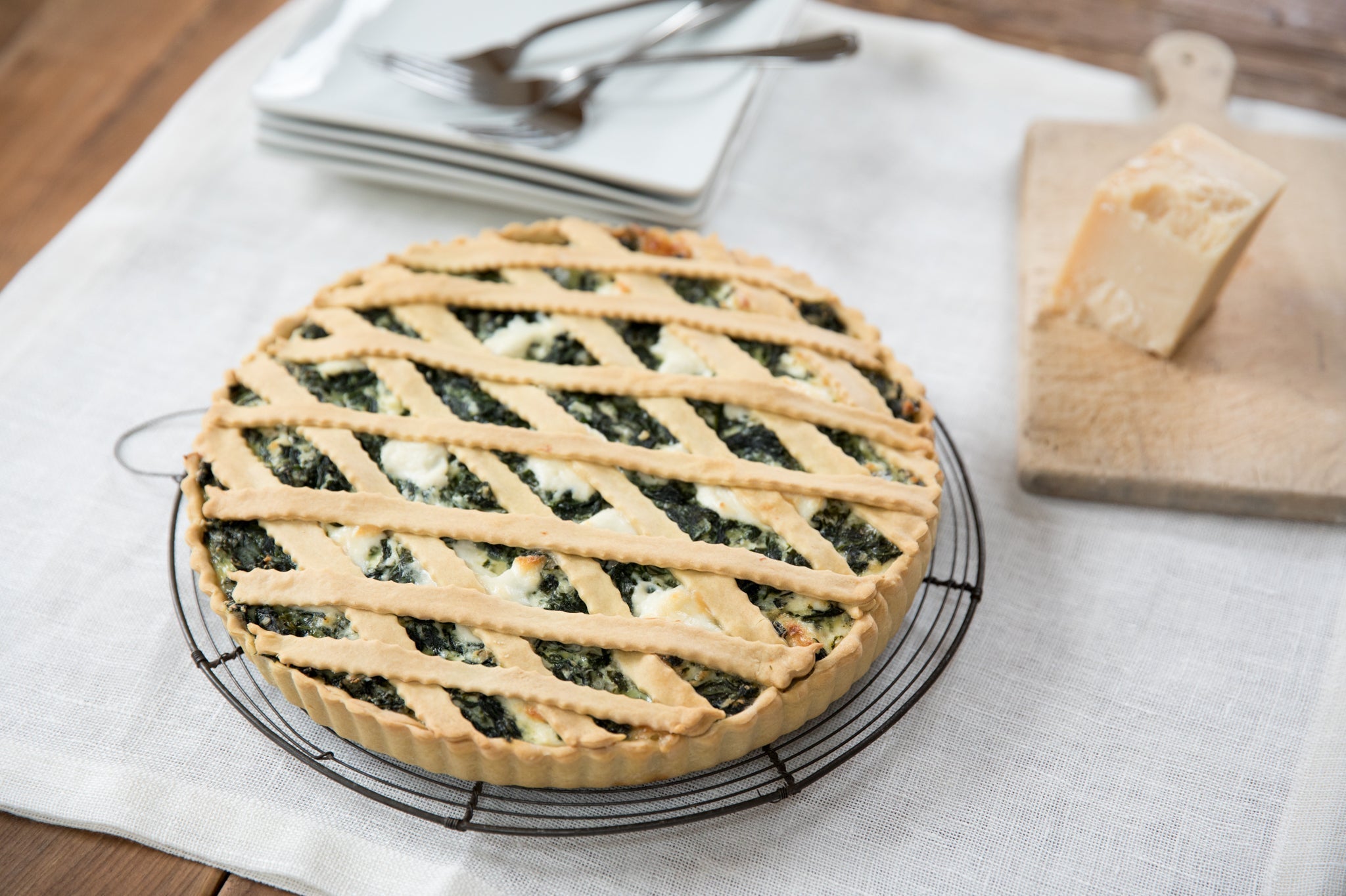 Einkorn spinach and ricotta pie cooling on a wire rack with a lattice crust, Parmesan wedge on a cutting board and stacked plates nearby