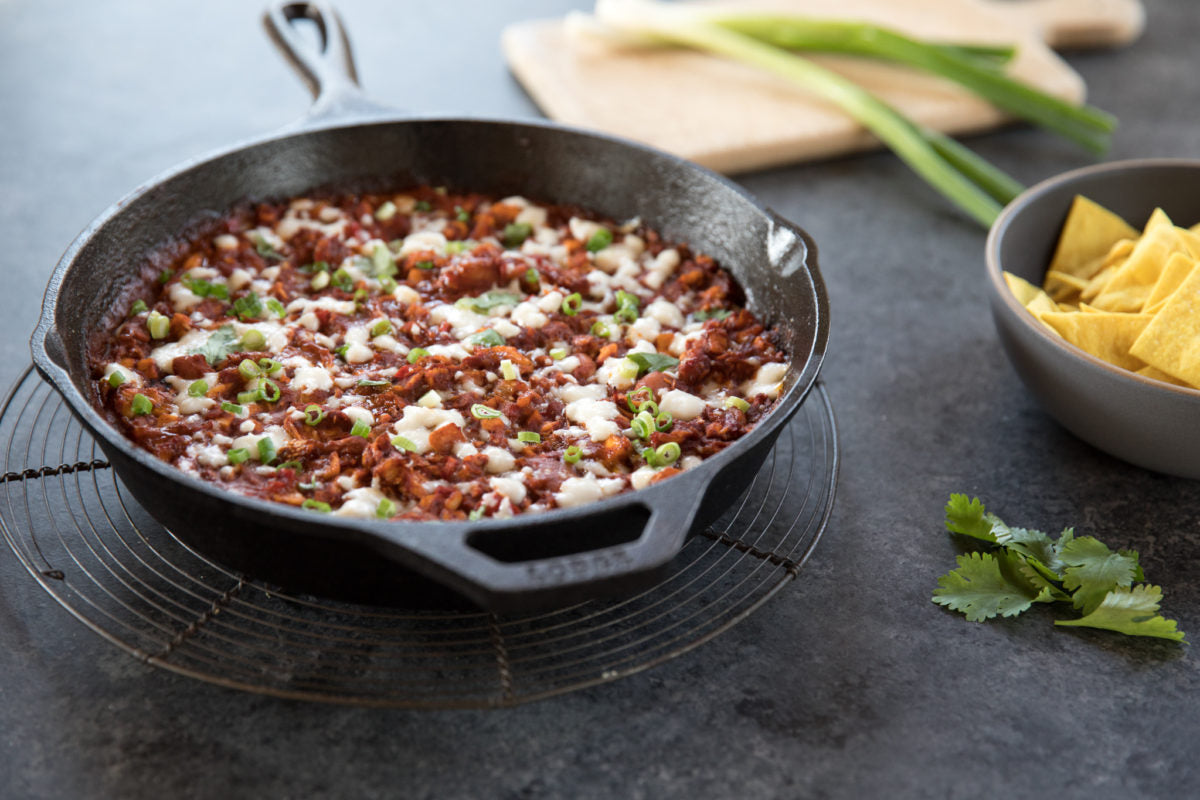 Angled view of skillet with chicken enchilada bean dip topped with melted cheese and scallions, tortilla chips at right