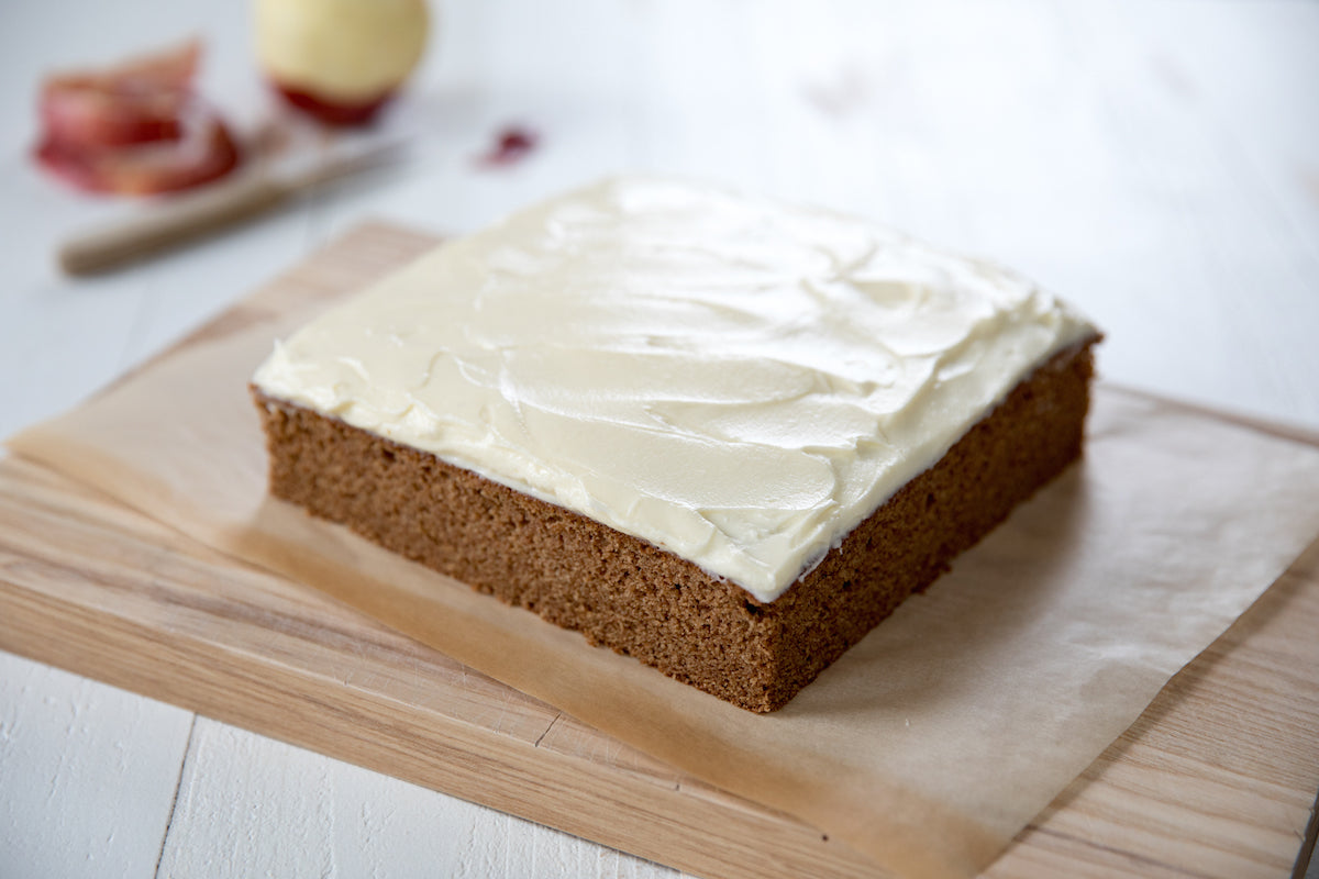 Three-quarter view of a square Einkorn applesauce spice cake topped with cream cheese frosting on parchment on a wooden cutting board