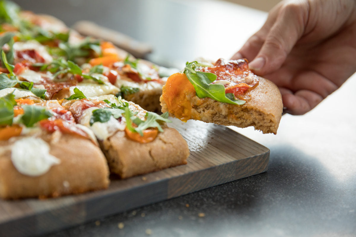 Close-up of sliced Einkorn whole-wheat focaccia on a cutting board with cheese, tomatoes, arugula and a hand lifting a slice