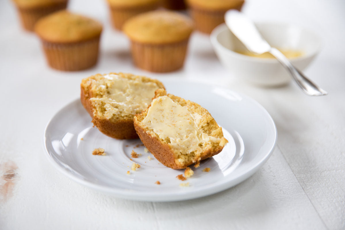 Halved Einkorn corn muffin spread with butter on a white plate, more muffins blurred in the background on a white table