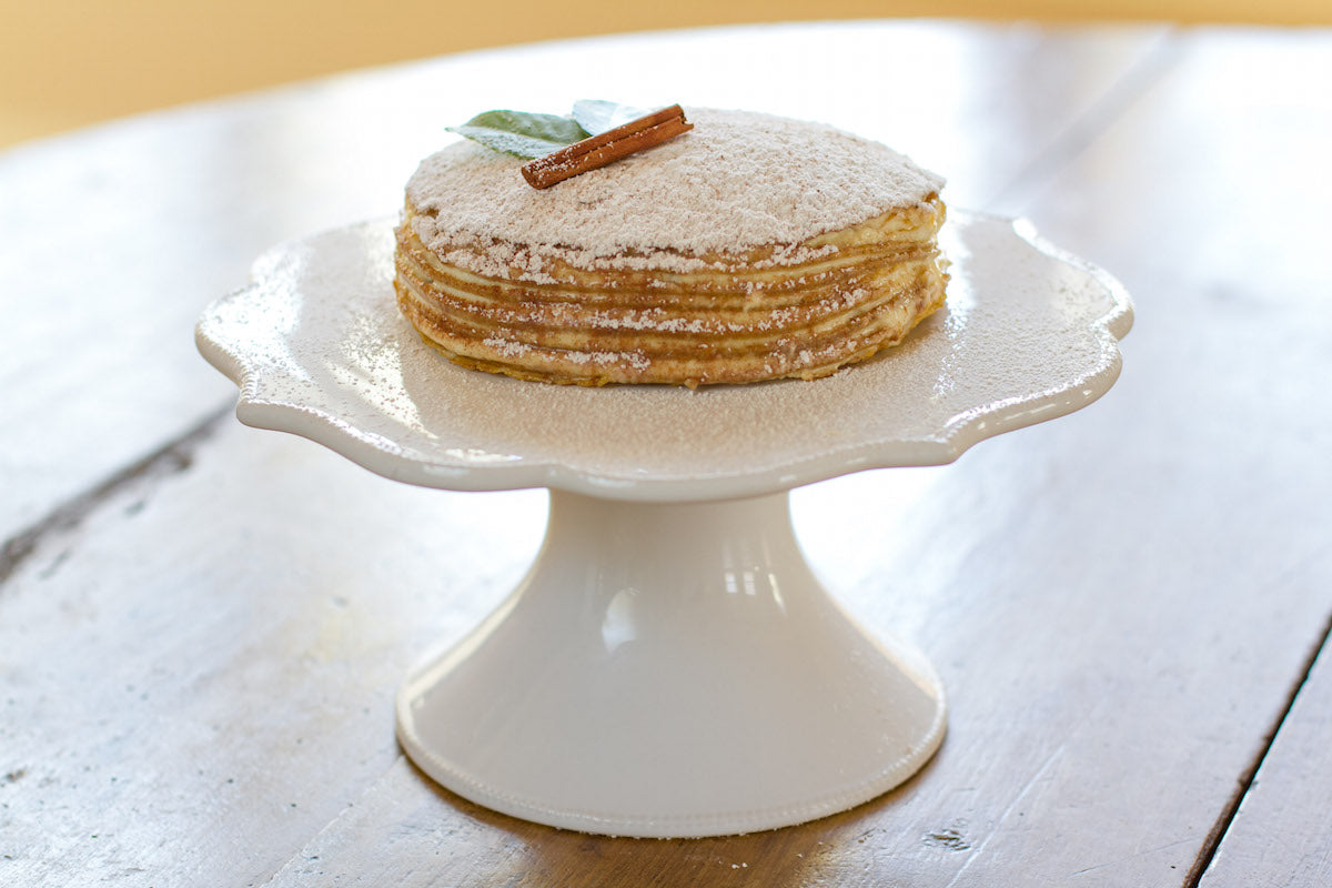 Front view of Einkorn pumpkin crepe mascarpone cake with stacked thin crepes, powdered sugar, cinnamon stick and leaf on a white cake stand