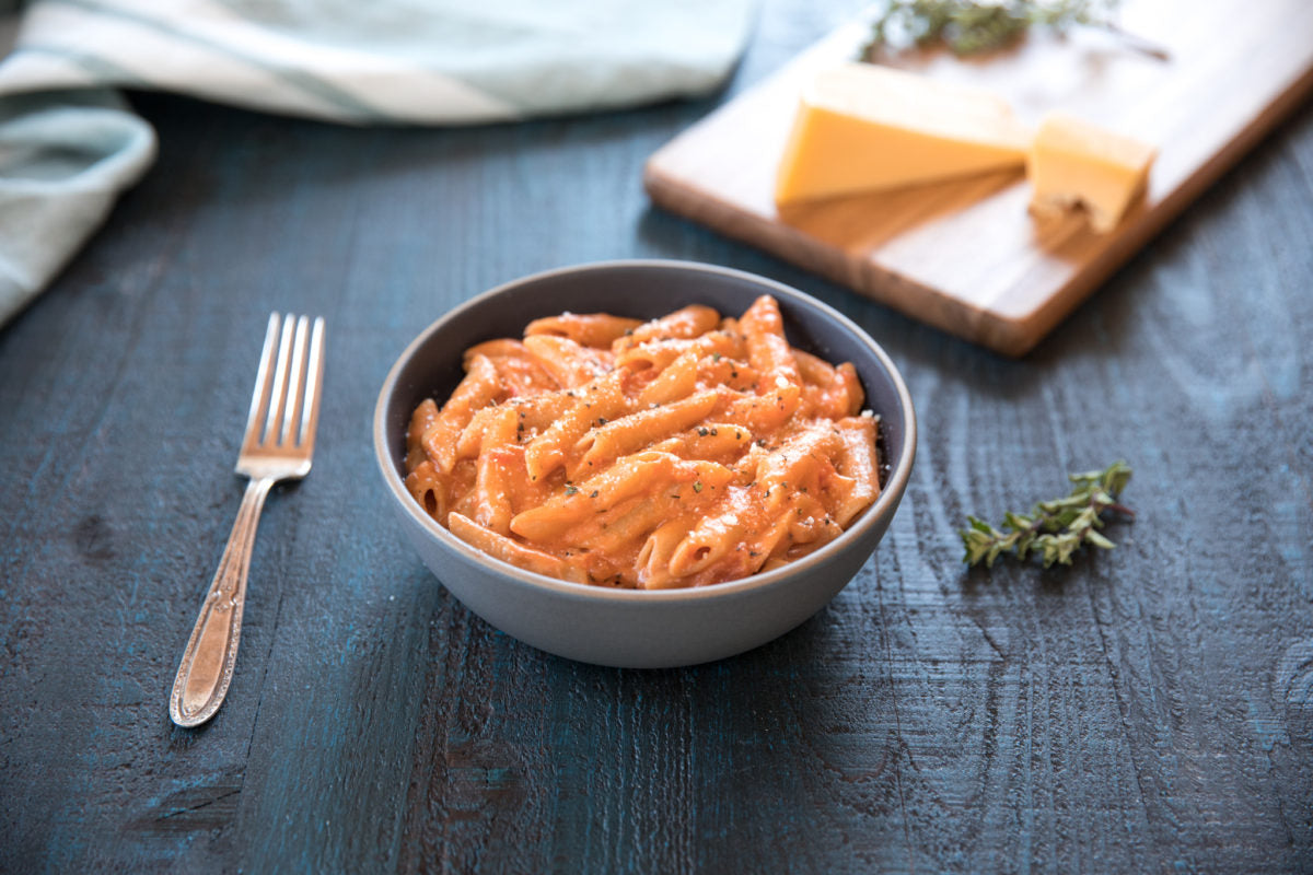 Bowl of penne mac and cheese in creamy tomato-cheese sauce on a dark wood table with a fork and a cheese board in the background