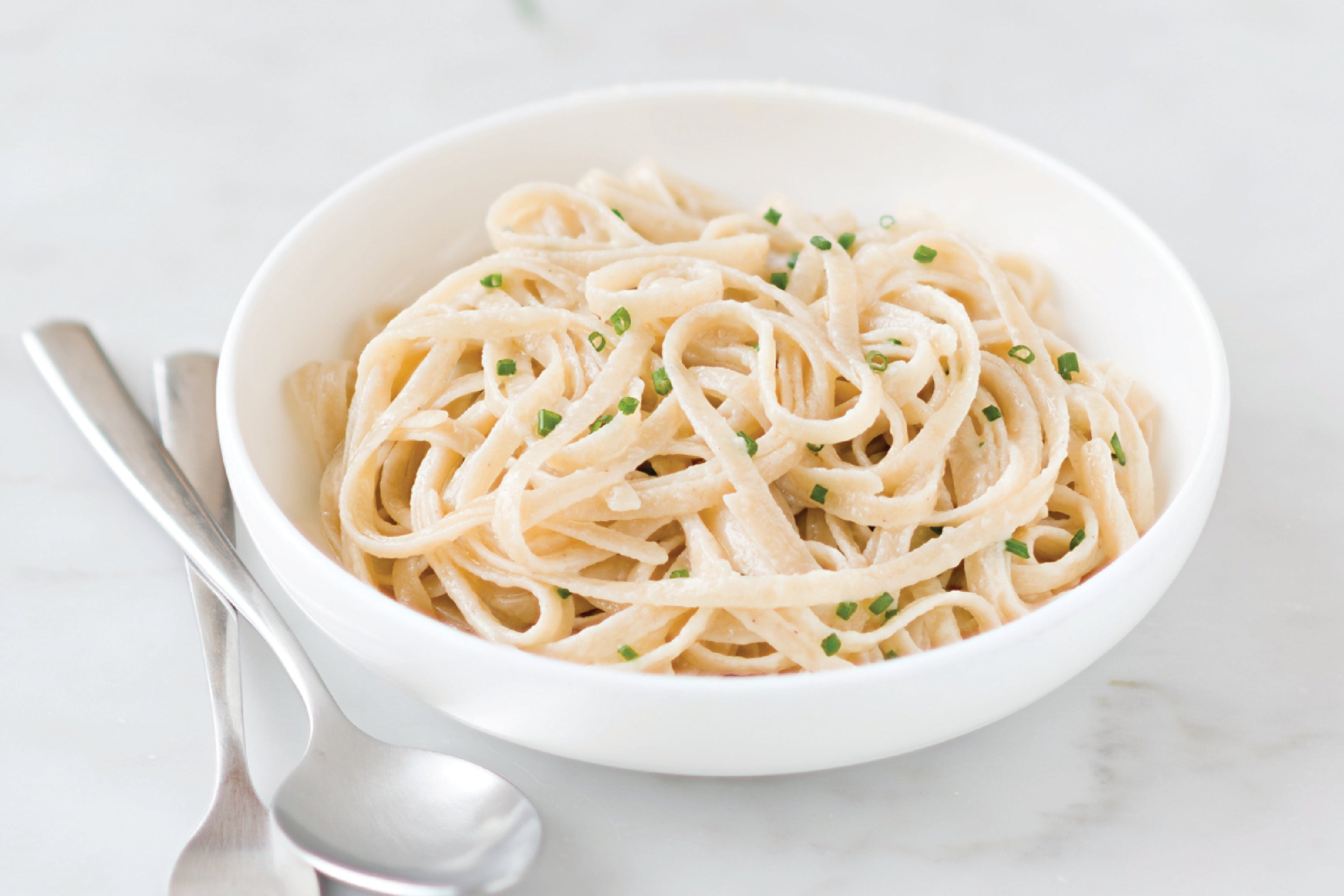 Bowl of Jovial brown rice fettuccine in creamy Alfredo sauce garnished with chives, on a marble surface with spoon and fork