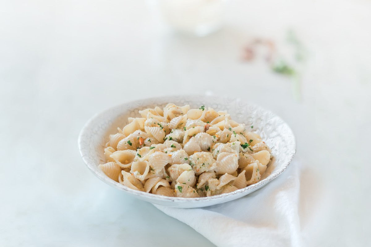 Bowl of gluten-free brown rice pasta shells tossed in creamy walnut sauce, garnished with parsley on a light tabletop