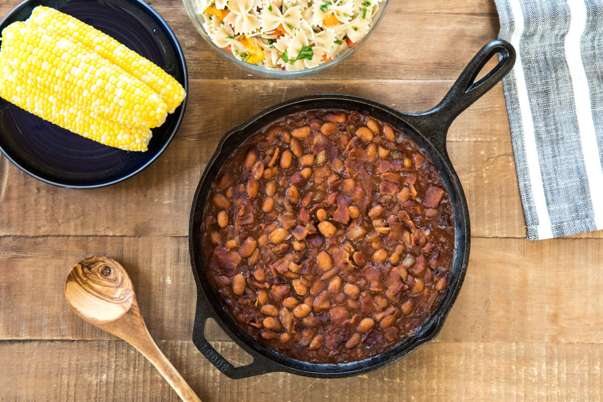 Baked borlotti beans in a cast-iron skillet on a wooden table with corn on the cob, a bowl of pasta salad, and a wooden spoon