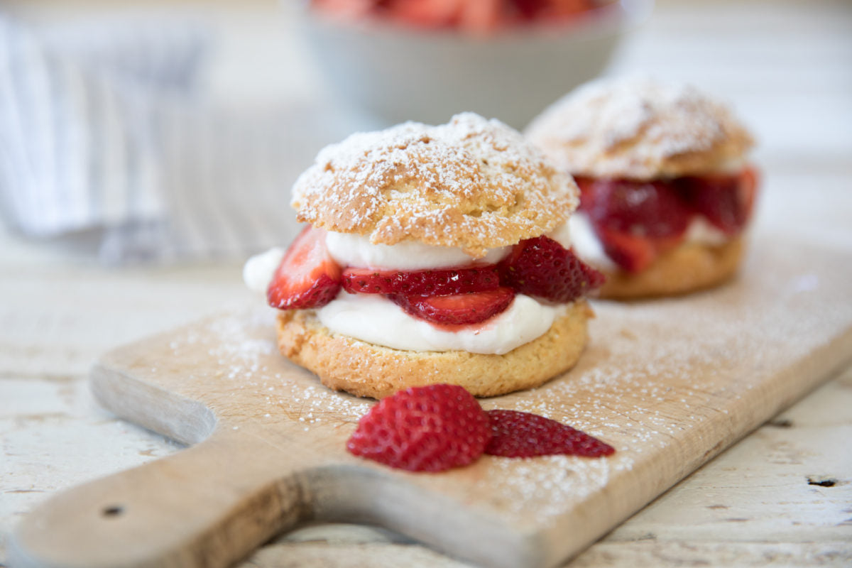 Two einkorn strawberry shortcakes layered with whipped cream and sliced strawberries on a wooden cutting board