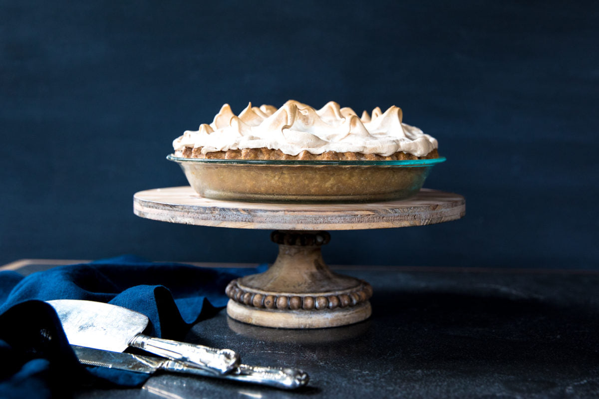 Side view of lemon meringue pie with browned meringue peaks and golden einkorn crust on a wooden cake stand against a dark blue backdrop