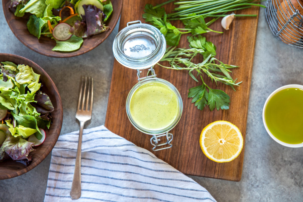 Jar of green goddess salad dressing on a wooden board with lemon half, fresh herbs, and two bowls of mixed salad