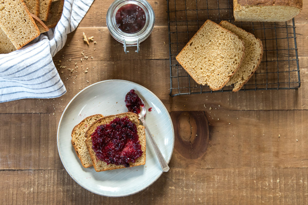 Two slices of Einkorn whole wheat sandwich bread spread with berry jam on a white plate, spoon, jar of jam and extra bread slices on a wooden table
