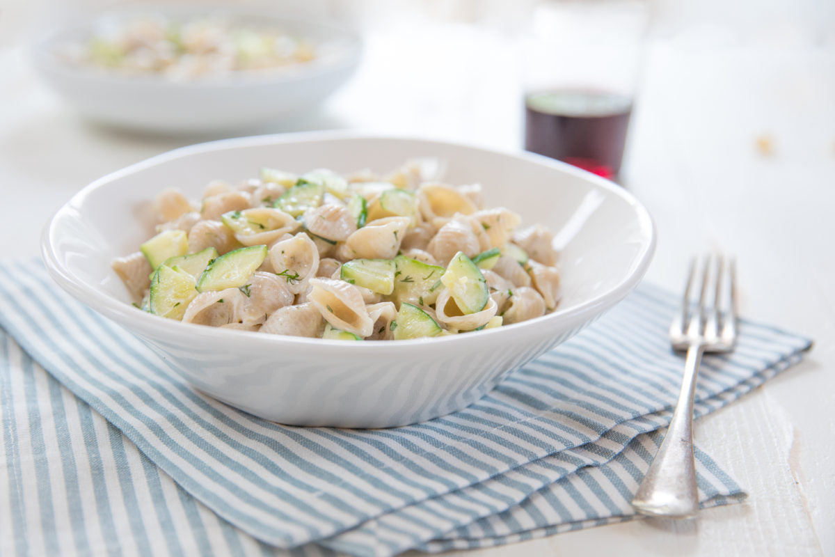 Bowl of Jovial gluten-free brown rice shell pasta tossed in lemon-herb Greek yogurt and dill with sautéed zucchini on a striped napkin