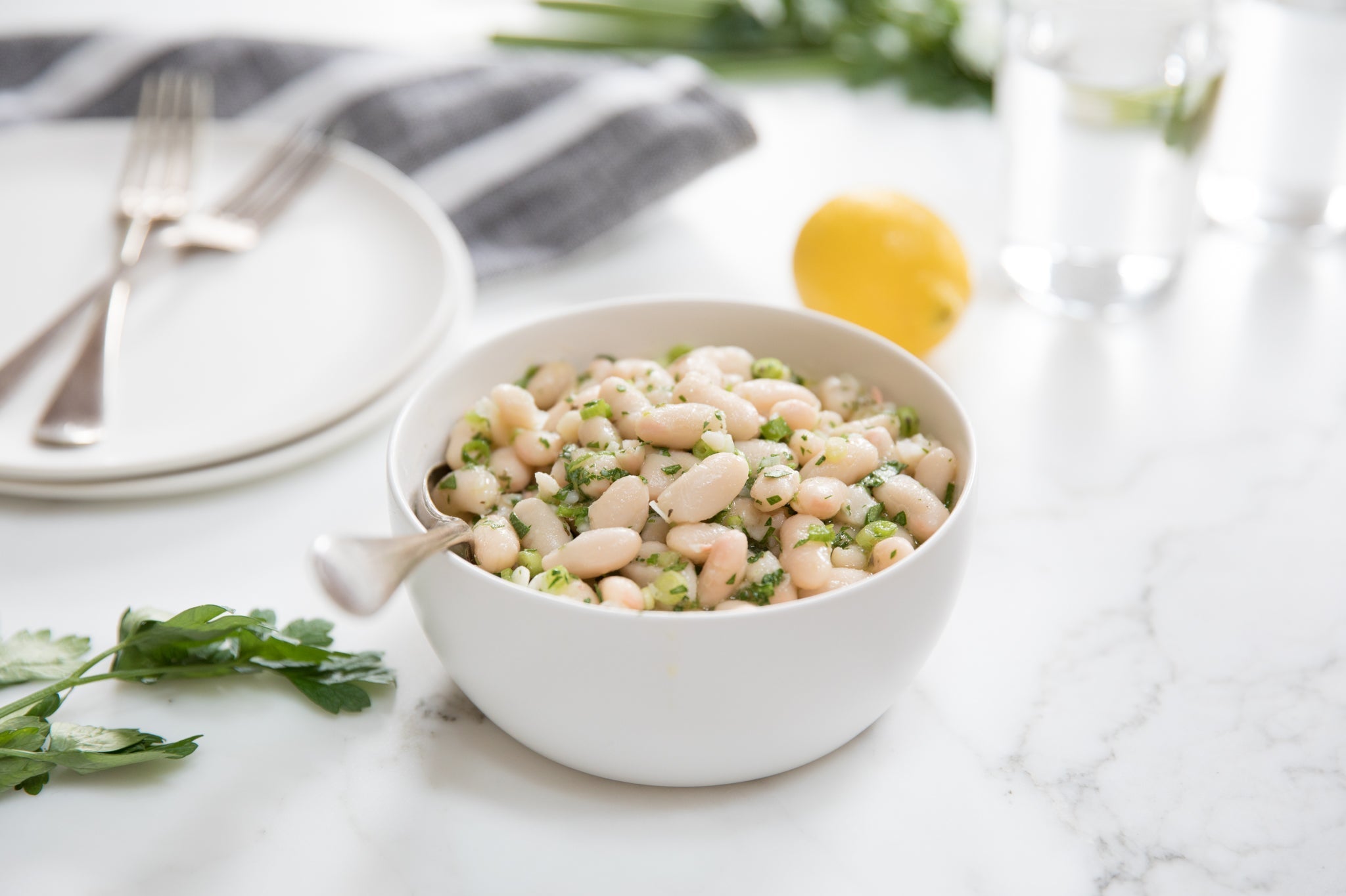 Bowl of Italian cannellini bean salad tossed with herbs and olive oil on a marble tabletop with lemon, parsley, and a fork