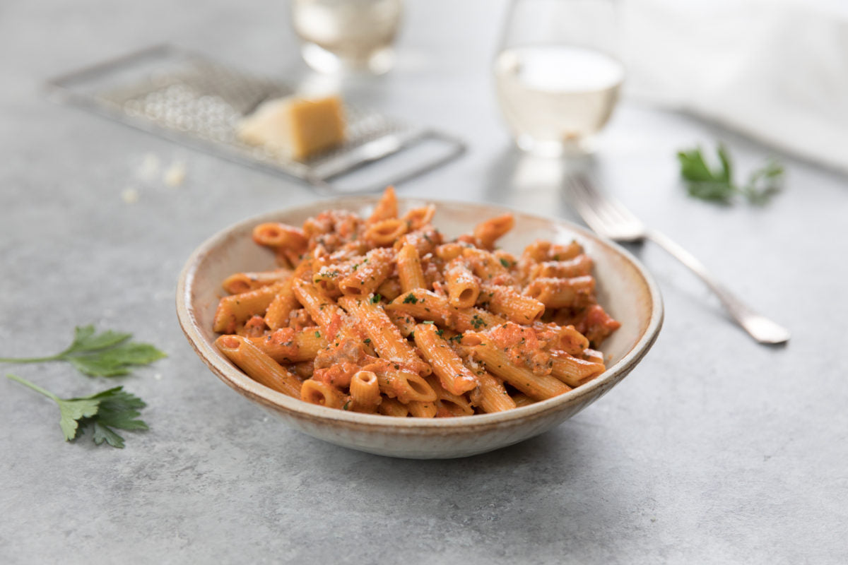 Bowl of penne and sausage in a creamy tomato sauce garnished with parsley, on a gray tabletop with a fork and cheese grater in soft-focus background